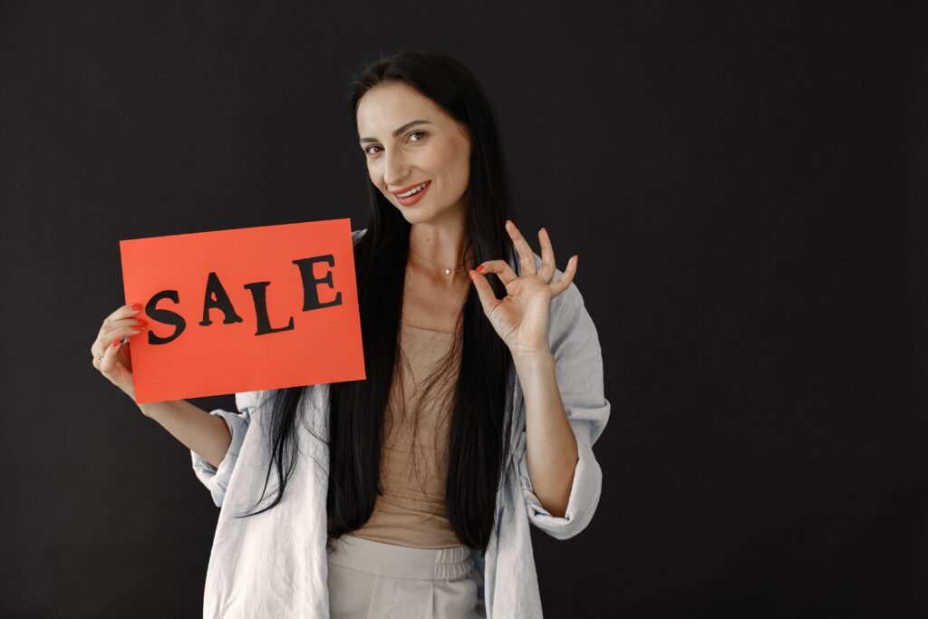 Brunette woman gesturing okay while holding a red sale sign on black background, perfect for consumerism and shopping themes.